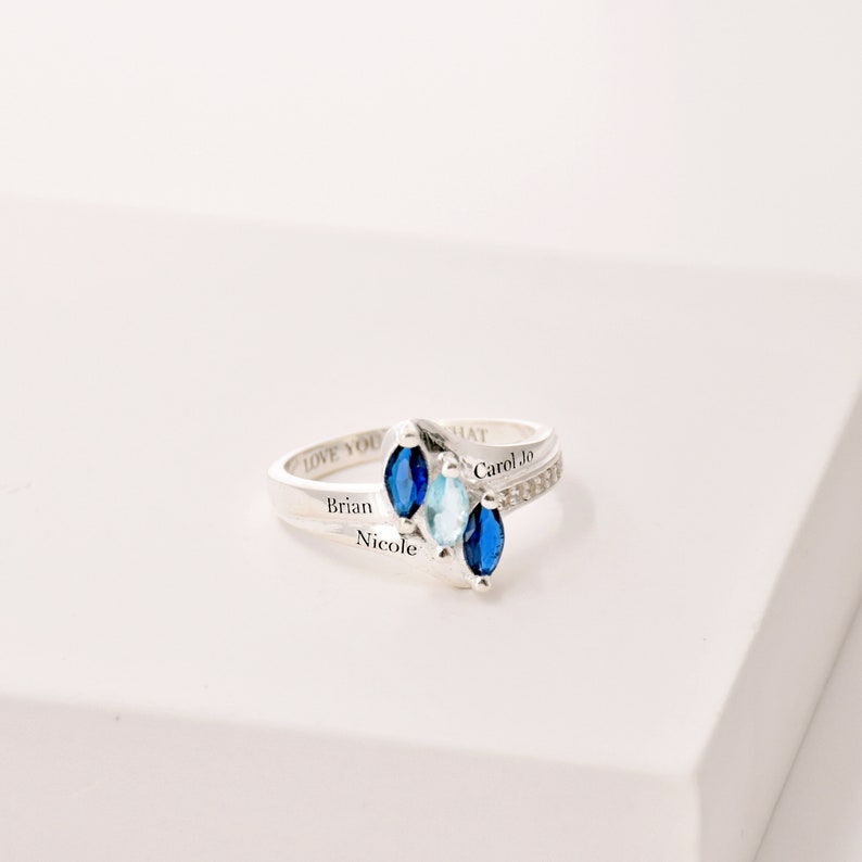 May include: A silver ring with three marquise-cut gemstones in shades of blue and aquamarine. The ring is engraved with the names "Brian", "Nicole", "Carol Jo", and the words "LOVE YOU" and "THAT".