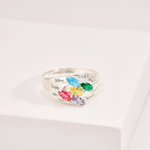 May include: A silver ring with a cluster of colorful gemstones. The ring features a band with engraved names and a design of blue, green, yellow, red, and purple oval-cut stones. The ring is on a white surface.