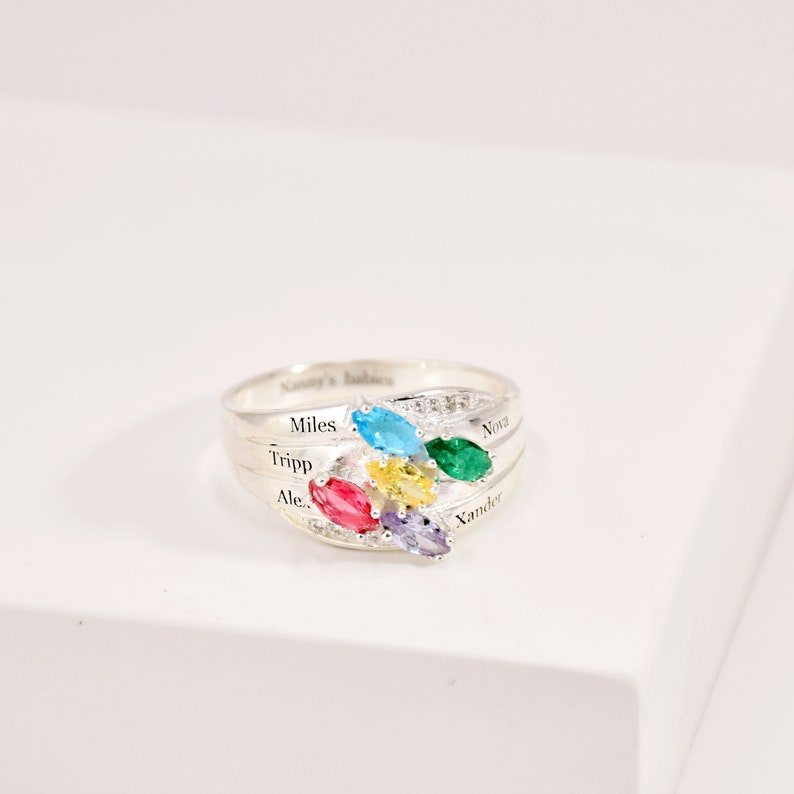 May include: A silver ring with a cluster of colorful gemstones. The ring features a band with engraved names and a design of blue, green, yellow, red, and purple oval-cut stones. The ring is on a white surface.