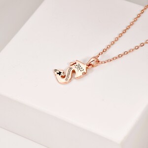 May include: Rose gold necklace with a graduation cap charm. The charm is engraved with the name "Romina" and the year "2022".