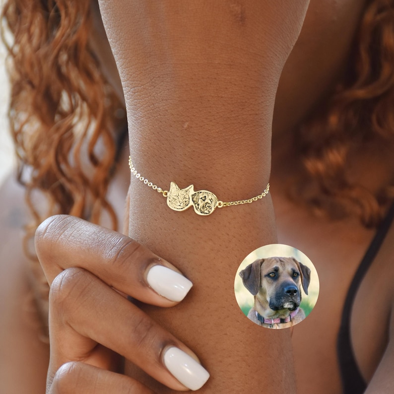 May include: A gold chain bracelet with two round charms featuring engraved images of a cat and a dog. The bracelet is worn on a person's wrist.