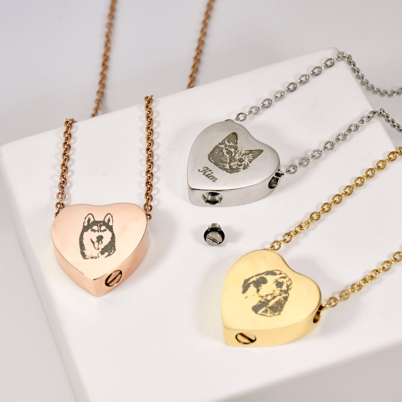 May include: Heart-shaped pet memorial necklaces in rose gold, silver, and gold. Each pendant features an engraved pet portrait and a screw-top opening. The necklaces are displayed on a white surface.