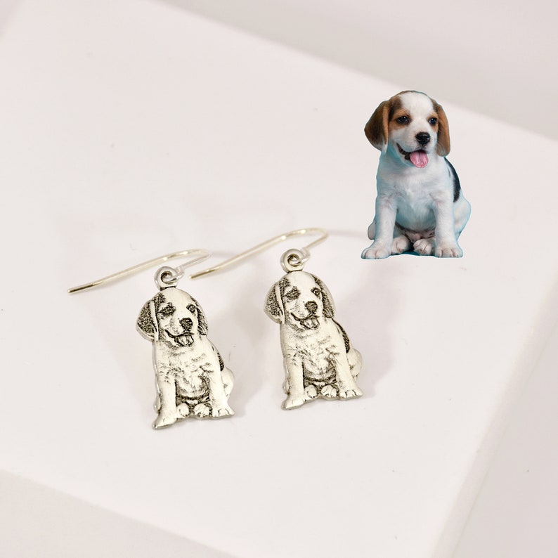 K&ouml;nnte beinhalten: Ein Paar silberne Ohrh&auml;nger mit einem kleinen, detaillierten Bild eines Beagle-Welpen. Der Welpe sitzt mit geneigtem Kopf und herausgestreckter Zunge.
