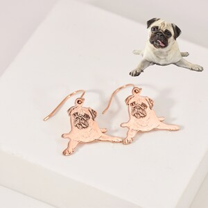 K&ouml;nnte beinhalten: Ohrringe aus ros&eacute;gold mit einem Anh&auml;nger in Form einer Mops-Silhouette. Die Ohrringe sind aus Metall und haben eine gl&auml;nzende Oberfl&auml;che.