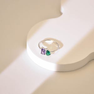 May include: A silver ring with a square purple gemstone and a teardrop green gemstone.
