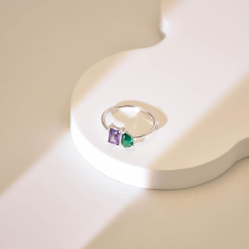 May include: A silver ring with a square purple gemstone and a teardrop green gemstone.