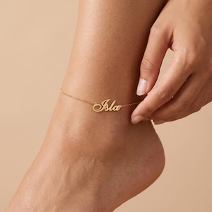 May include: A gold-toned anklet featuring a delicate chain and a central nameplate pendant with the name Isla written in a cursive script font. The piece is designed with a thin metal link chain that secures around the ankle with a clasp.