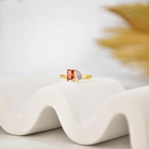 Pu&ograve; includere: Un anello d'oro con una gemma rossa rettangolare e una gemma viola a forma di pera. L'anello &egrave; posto su una superficie bianca ondulata, con uno sfondo sfocato.