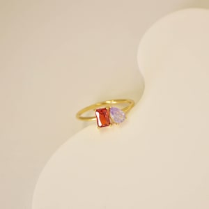 Pu&ograve; includere: Un anello d'oro con una gemma rossa rettangolare e una gemma viola a forma di pera. L'anello ha una fascia sottile. Le pietre preziose sono il punto focale, mostrando i loro colori vivaci e le forme uniche. L'anello &egrave; su uno sfondo morbido e neutro.
