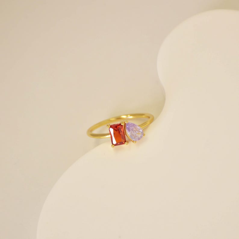 Pu&ograve; includere: Un anello d'oro con una gemma rossa rettangolare e una gemma viola a forma di pera. L'anello ha una fascia sottile. Le pietre preziose sono il punto focale, mostrando i loro colori vivaci e le forme uniche. L'anello &egrave; su uno sfondo morbido e neutro.