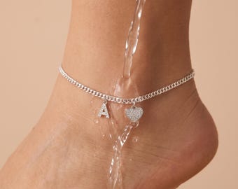 May include: A silver-toned chain anklet featuring two dangling charms: a letter A pendant and a heart-shaped pendant, both encrusted with small clear stones. The anklet is shown with water flowing over it, highlighting the metallic finish and design.
