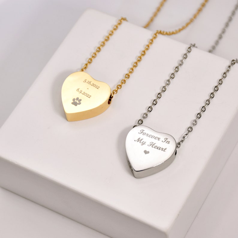 May include: Two heart-shaped pendant necklaces. One is gold-toned with engraved dates and a paw print. The other is silver-toned with the inscription "Forever In My Heart" and a small heart symbol. Both have delicate chain necklaces.