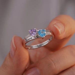 May include: A silver ring featuring two heart-shaped gemstones, one purple and one blue, with small clear stones. The ring is engraved with the names "Diane" and "David", and the phrase "MY LOVE FOREVER X".