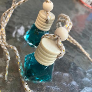 May include: Two small, square glass bottles filled with teal liquid, each with a wooden cap and a decorative rope. The rope is beige with gold accents. The bottles are hanging, suggesting they are car air fresheners.