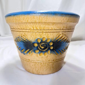 May include: A tan-colored ceramic flower pot with a textured surface. The rim is painted in a solid blue. A central embossed floral design features a dark blue center surrounded by yellow petals and blue leaf-like accents on each side.
