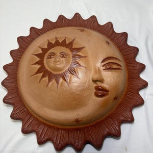 May include: A circular terracotta wall plaque featuring a relief design of a sun and moon face. The sun is depicted with rays and a smiling face, while the moon is shown with a closed eye and lips. Small star shapes are etched into the surface.