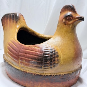 May include: A ceramic planter shaped like a sitting hen with a yellow body, brown wing details, and a dark brown base. The hollow center is open at the top, and the surface features a textured, matte finish with blended earth-tone colors.