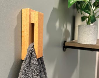 May include: A wooden towel hook mounted on a gray wall. The hook is made of light-colored wood and holds a gray towel. A shelf with a potted plant is visible in the background, adding a touch of greenery to the scene.