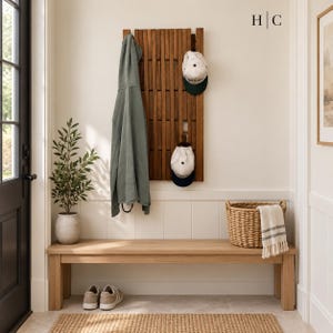 May include: A vertical wooden wall-mounted coat rack with slats, holding a sage green hoodie and two baseball caps. Below is a long wooden bench, a woven basket with a striped cloth, and a pair of sneakers. The logo H|C is visible at the top.