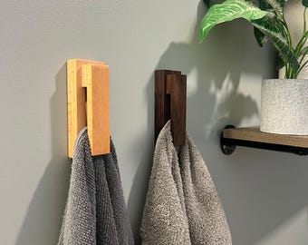 May include: Two wooden towel hooks, one light and one dark, mounted on a gray wall. Gray towels hang from the hooks. A shelf with a plant is visible in the background, adding a touch of greenery to the bathroom setting.