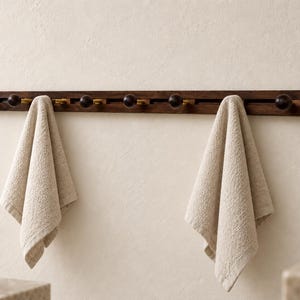 May include: A horizontal dark wood wall-mounted rack featuring a series of round knobs on brass-colored stems. Two beige textured towels are draped over the knobs. The rack has a linear design with multiple pegs for hanging items.