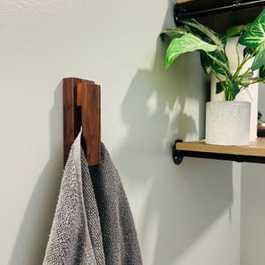 May include: A dark wood towel hook mounted on a light green wall, holding a gray towel. The hook has a rectangular design with a slot for the towel. A shelf with a plant is visible in the background.