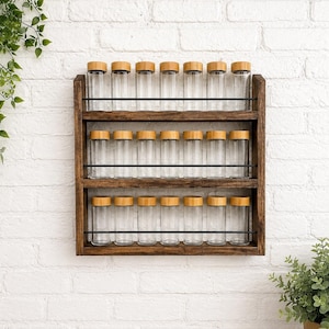May include: A wooden spice rack with three shelves, each holding clear glass jars with bamboo lids. The rack is mounted on a white brick wall, with greenery in the background. The rack is designed to hold 18 spice jars.
