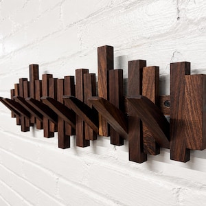 May include: A wall-mounted coat rack constructed from dark brown wood, featuring a series of vertical rectangular slats of varying heights and several angled hooks extending outward for hanging items, showcasing a natural wood grain texture.