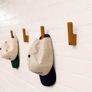 May include: Four L-shaped wooden wall hooks mounted on a white brick wall. Two baseball caps are hanging from the hooks: one cream cap with a dark green brim and one cream cap with a navy blue brim, both featuring black text on the front.