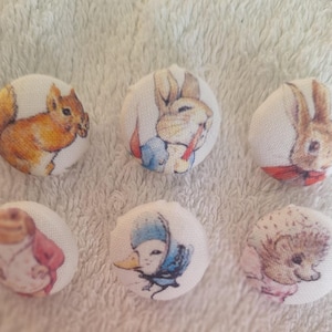 May include: Six round fabric-covered buttons featuring printed illustrations of woodland animals. The designs include a squirrel, a rabbit in a blue jacket, a rabbit with a red bow, a frog in a red coat, a duck in a blue bonnet, and a hedgehog.