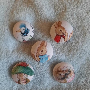 May include: Five round fabric buttons featuring characters from the children's book, Peter Rabbit. The buttons feature Peter Rabbit, Benjamin Bunny,  a hedgehog, a bird, and a rabbit wearing a green hat.