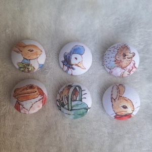 May include: Six circular fabric buttons with illustrations of storybook characters. The buttons show a rabbit, duck, hedgehog, frog, and other animals. The buttons are white with colourful illustrations.