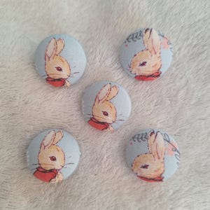 May include: Five round fabric buttons with a light blue background and a printed image of a brown rabbit with red accents. The buttons are ideal for adding a touch of whimsy to any craft project.