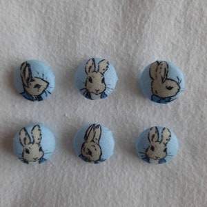 May include: Six light blue fabric-covered buttons featuring Peter Rabbit illustrations. Each button shows a different image of the rabbit's face, set against a light blue background. The rabbit is white and beige.