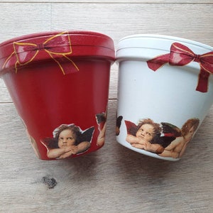 May include: Two terracotta flower pots, one red and one white, decorated with cherub angels and red bows.