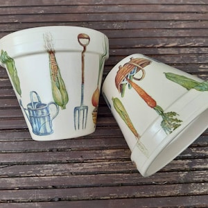 May include: Two white ceramic flower pots with a garden theme. The pots are decorated with illustrations of vegetables, gardening tools, and a watering can. The pots are on a wooden surface.