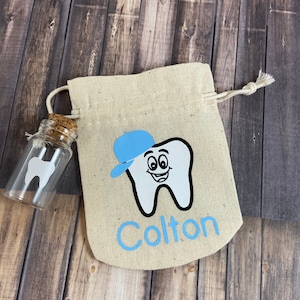May include: A small white canvas bag with a blue tooth design and the name "Colton" embroidered on it. The bag is tied with a string and has a small glass bottle with a cork stopper and a white tooth design inside.