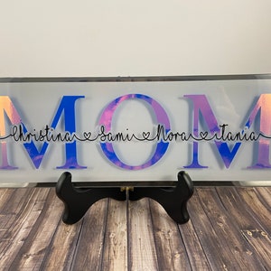 May include: A white rectangular glass sign with the word "MOM" in large iridescent letters. The letters have a purple and blue ombre effect. Below the letters, the names "Christina", "Samir", "Nora", and "Ivania" are written in black cursive with hearts between each name.
