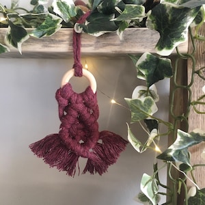 May include: A burgundy macrame wall hanging with a wooden ring and tassels. The hanging is made with a thick, soft cord and has a bohemian style.