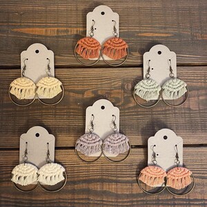 May include: Set of six pairs of handmade macrame hoop earrings. Each pair features a different color of macrame, including orange, yellow, green, beige, and pink. The earrings are displayed on white card stock.