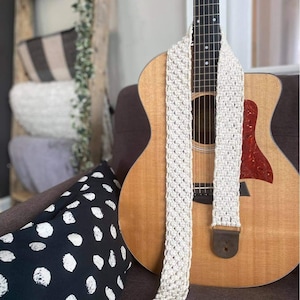Puede incluir: Una correa de guitarra de crochet blanco con extremos de cuero marrón. La correa está colgada de una guitarra acústica con un acabado de madera natural.