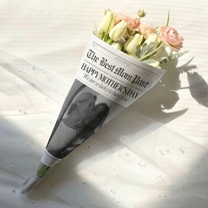 May include: A bouquet of flowers wrapped in newspaper with the text "The Best Mom Post HAPPY MOTHER'S DAY". The bouquet includes white tulips and pink roses. The newspaper wrap features a black and white photo of two people.