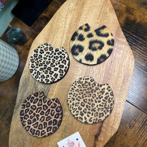 May include: Four round car coasters with a leopard print design in shades of brown, tan, and black. The coasters are displayed on a wooden cutting board. A small white card with the text "Bella Phoenix Crystals" is also visible.