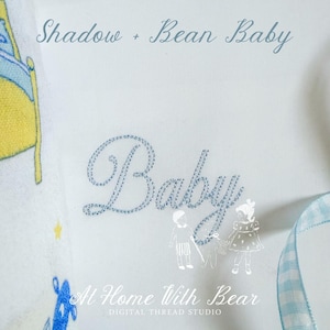 May include: White baby blanket with the word "Baby" embroidered in light blue script. The blanket also features the text "Shadow + Bean Baby" and "At Home With Bear". A light blue ribbon is visible.
