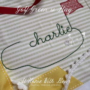以下が含まれることがあります： 緑と白のストライプ模様の生地アイテム。「Charlie」の文字が緑色の筆記体で刺繍されています。赤い星型の旗が上部に縫い付けられています。「Golf Green w Flag」の文字が上部にあります。