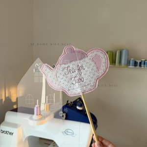 Puede incluir: Una tetera de tela rosa y blanca sobre un palo de madera. La tetera tiene bordadas las palabras "Tea for Two". El fondo incluye una máquina de coser y carretes de hilo.