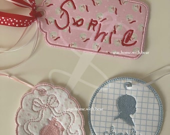Pode incluir: Três etiquetas de presente de tecido rosa e azul com desenhos bordados. A primeira etiqueta diz "Sophie" em bordado vermelho. A segunda etiqueta tem um desenho bordado rosa de uma menina com um laço no cabelo. A terceira etiqueta tem uma silhueta azul de uma pessoa com o nome "Charlie" bordado em branco.