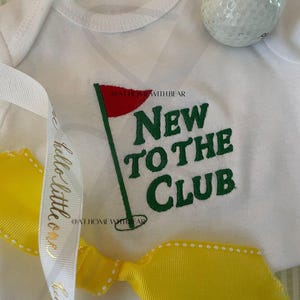 May include: White baby onesie with a green and red golf club graphic and the text "New to the Club". A yellow ribbon with white polka dots is tied around the onesie. A white golf ball is in the background.