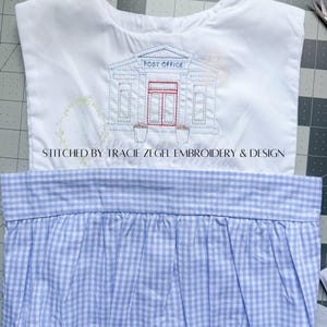 May include: White top with a blue and red embroidered post office design. The top is paired with a blue and white gingham skirt. The text "STITCHED BY TRACIE ZEGEL EMBROIDERY & DESIGN" is printed on the top.