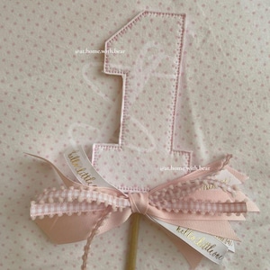 May include: Pink felt number one with pink and white ribbon bow and the text "hello little one" on the ribbon. The background is a pink polka dot fabric.